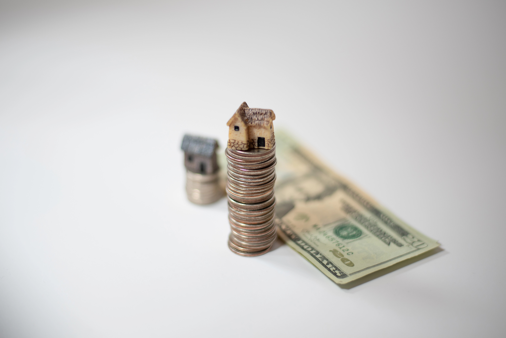 Co może być wkładem własnym – stos monet z miniaturowym domkiem na szczycie oraz banknoty dolarowe w tle, symbolizujące pieniądze przeznaczone na wkład własny przy zakupie nieruchomości.