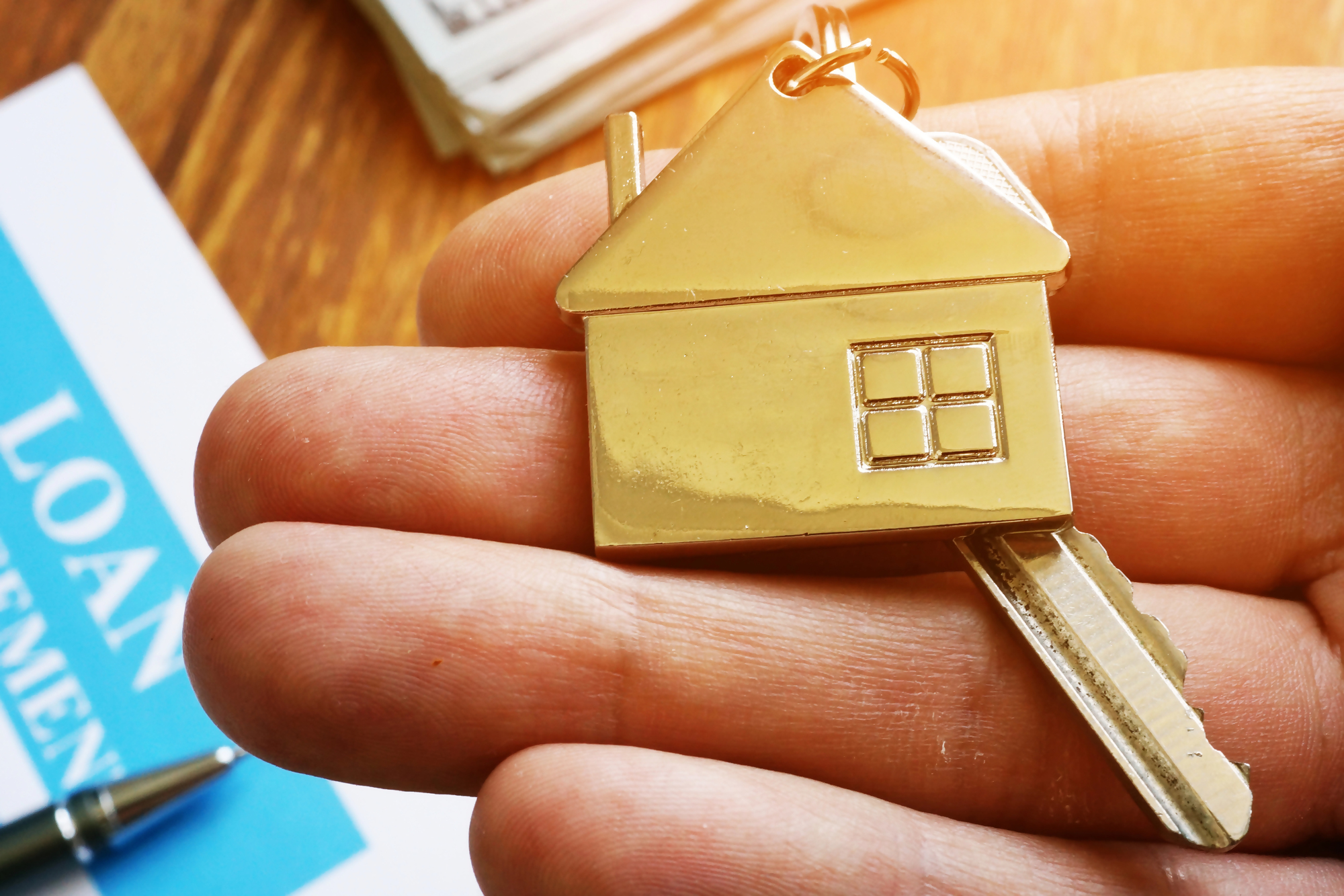 Kredyt hipoteczny krok po kroku – mosiężny brelok w kształcie domu z kluczem trzymany w dłoni, w tle dokumenty i długopis symbolizujące proces uzyskania kredytu hipotecznego.