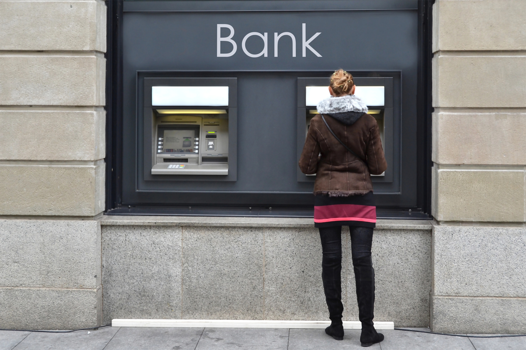 Kobieta stoi przy bankomacie przed budynkiem banku, obrazek ilustruje temat "gdzie wyrzucić kartę płatniczą" oraz przypomina o bezpiecznej utylizacji zużytej karty płatniczej.