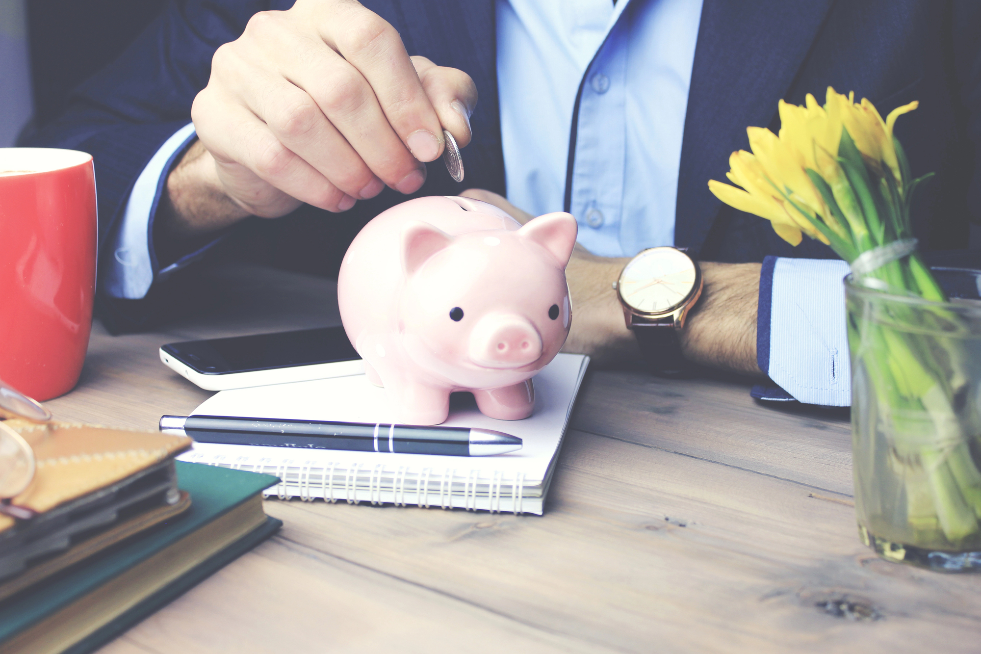 Intercyza – mężczyzna w garniturze wrzuca monetę do skarbonki w kształcie świnki, symbolizując oszczędzanie i zarządzanie majątkiem w kontekście intercyzy na biurku z notatnikiem oraz kwiatami.