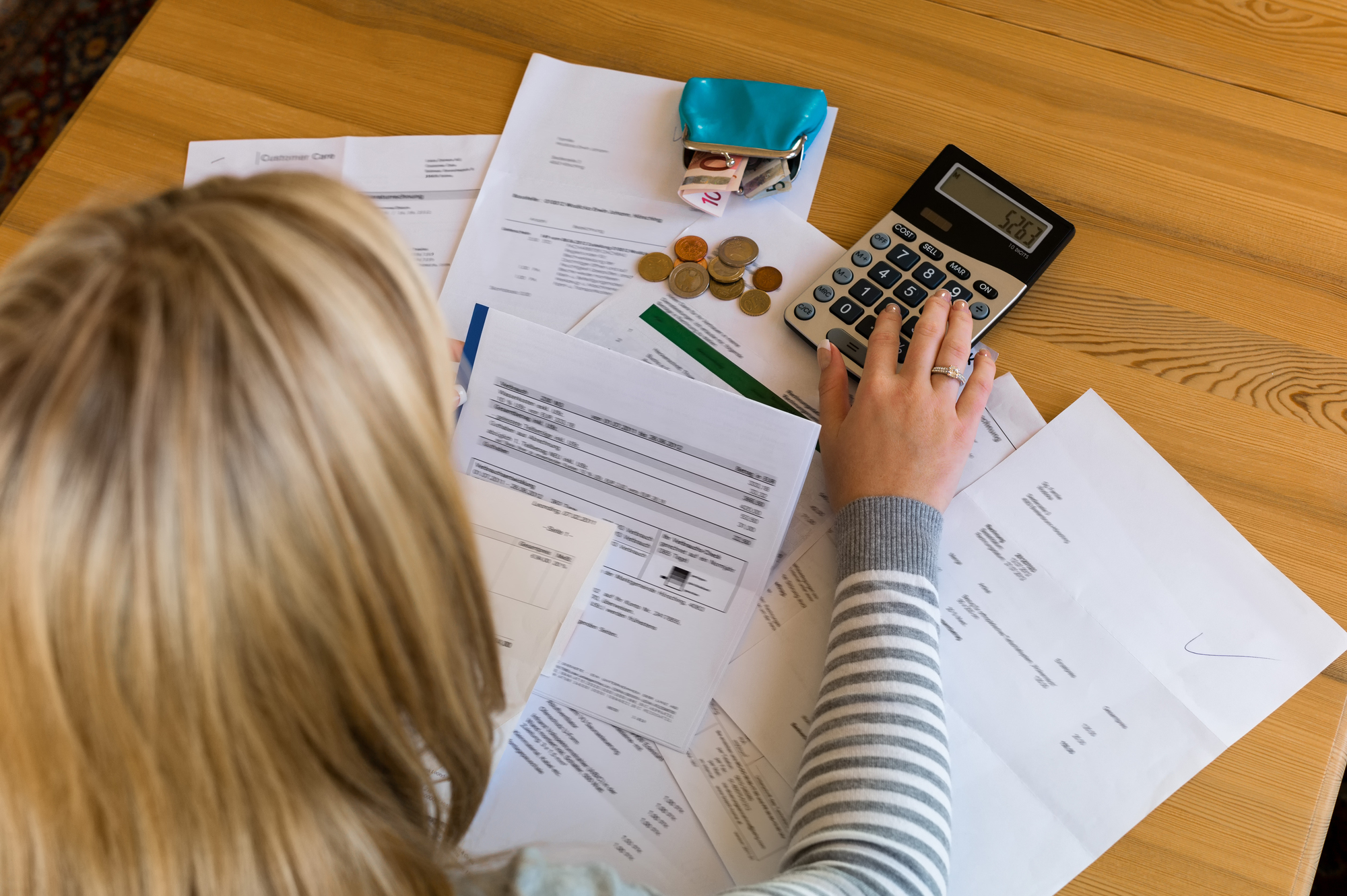 Kobieta przegląda dokumenty finansowe i wylicza wydatki na kalkulatorze, co ilustruje problem "brak ubezpieczenia mieszkania a kredyt hipoteczny" oraz związane z tym konsekwencje finansowe dla kredytobiorcy. Na stole widoczne są monety, portfel i liczne rachunki.