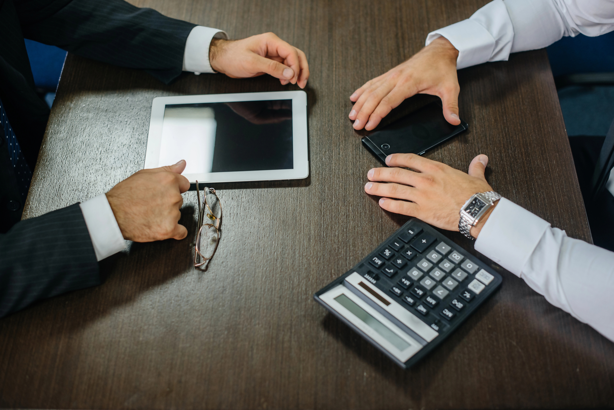 Dwie osoby w garniturach omawiają promesa kredytowa przy stole, na którym znajdują się kalkulator, tablet i telefon, ilustracja biznesowego spotkania dotyczącego kredytu