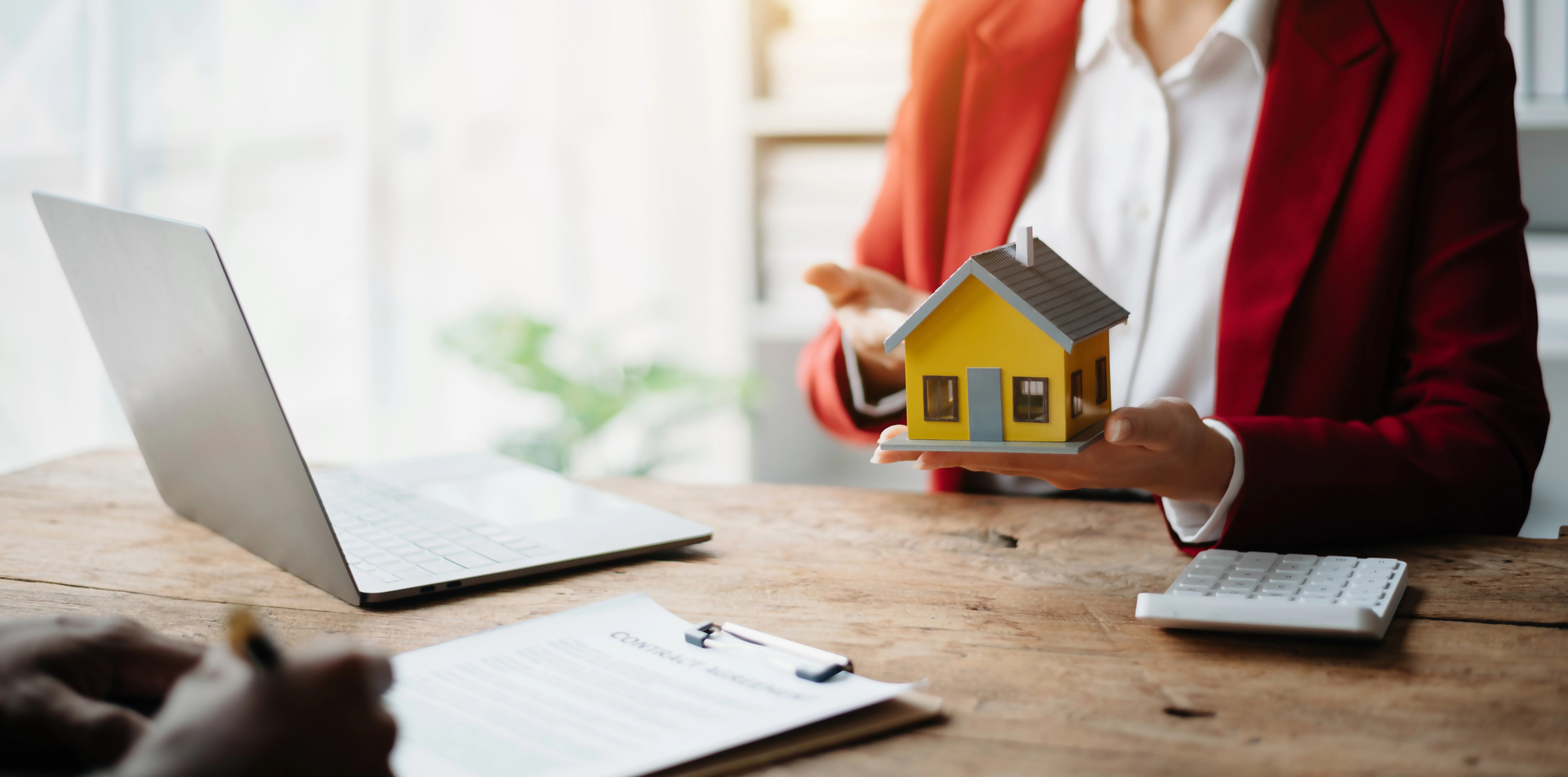 Kobieta w czerwonej marynarce omawia harmonogram spłaty kredytu hipotecznego, trzymając w ręku model domu, przy stole z laptopem, kalkulatorem i dokumentami.