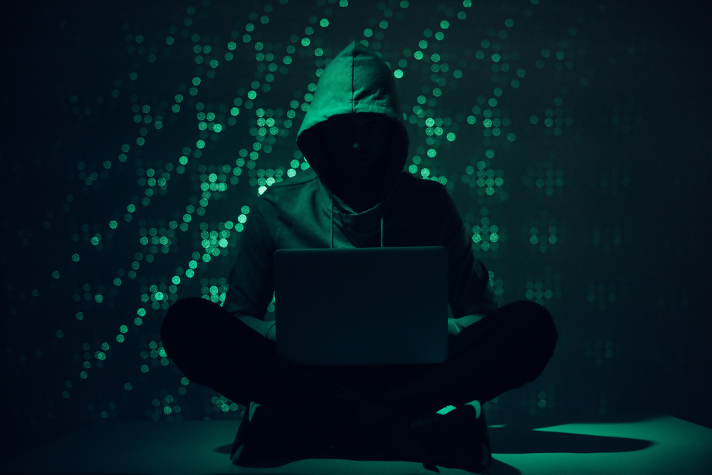 Osoba w kapturze siedząca w ciemnym pomieszczeniu przed laptopem, symbolizująca hakera lub cyberprzestępczość, tło w stylu cyfrowym z zielonymi światełkami.