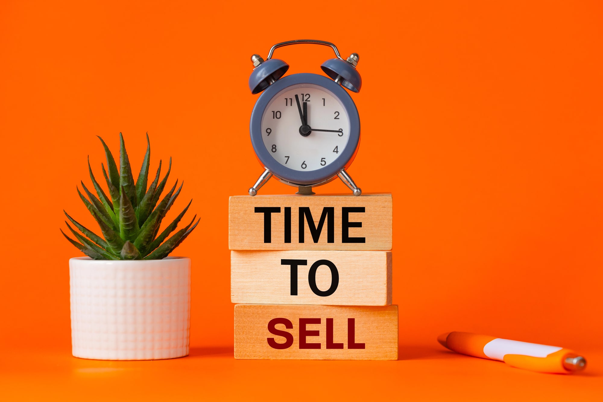 Na tle w intensywnym pomarańczowym kolorze znajdują się klocki z napisem "TIME TO SELL", obok zegar budzik i roślina doniczkowa. Obrazek symbolizuje moment, w którym warto rozważyć kwestie takie jak cena emisyjna na rynku.