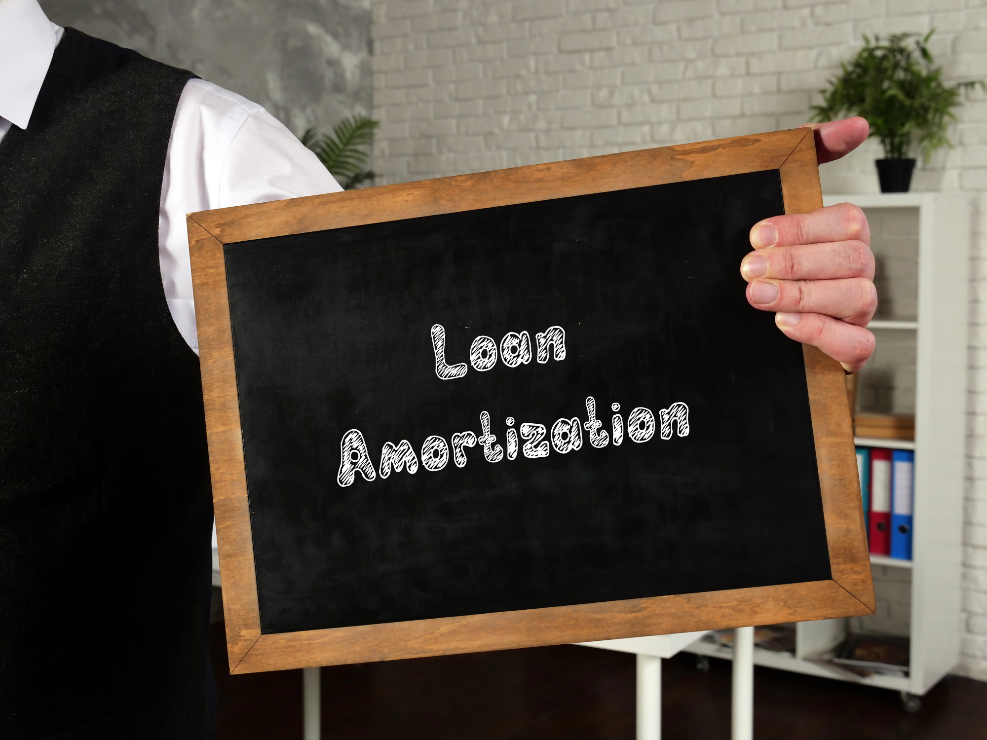 Osoba trzymająca tabliczkę z napisem "Loan Amortization", co odnosi się do procesu jakim jest amortyzacja kredytu, przedstawione w biurowym otoczeniu.