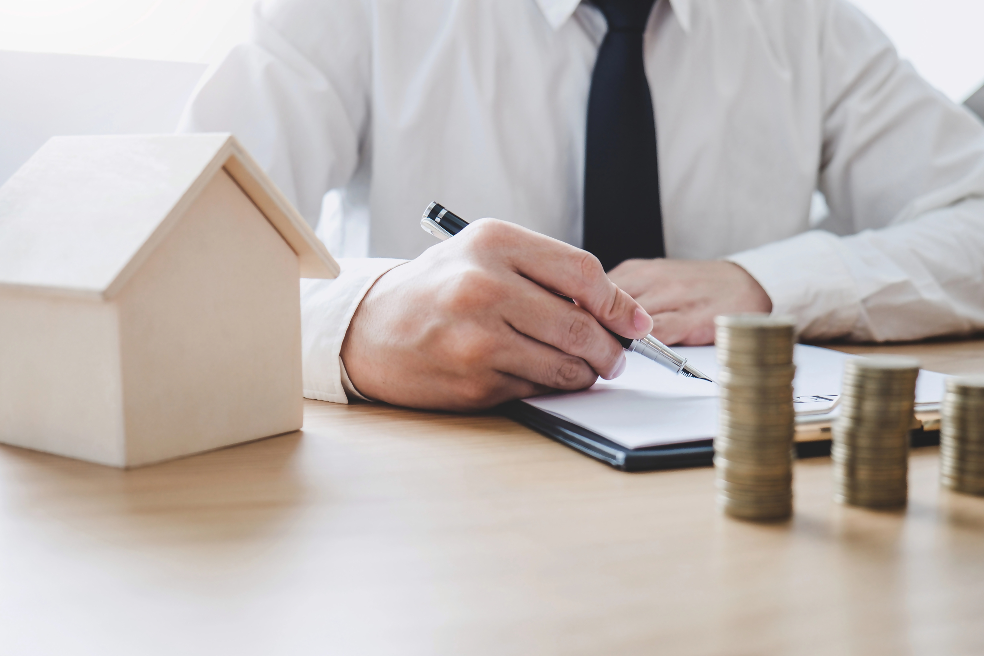 Ile się czeka na kredyt hipoteczny – mężczyzna w garniturze podpisuje dokumenty związane z kredytem hipotecznym, obok stoi model domu i stosy monet, symbolizujące finanse i zakup nieruchomości.