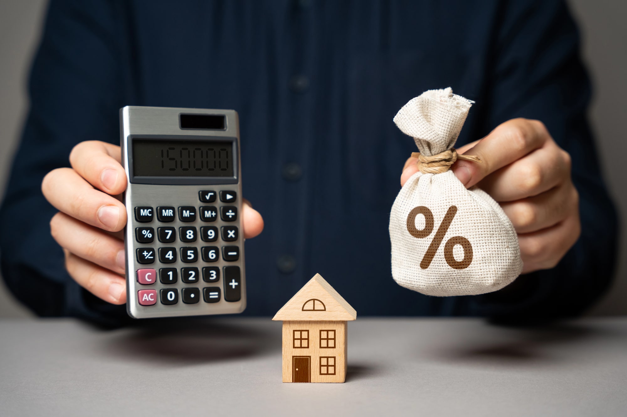 Osoba trzymająca kalkulator z kwotą 150000 oraz worek na pieniądze z symbolem procentów, obok drewniany model domu. Tematyka związana z cashback i finansami.