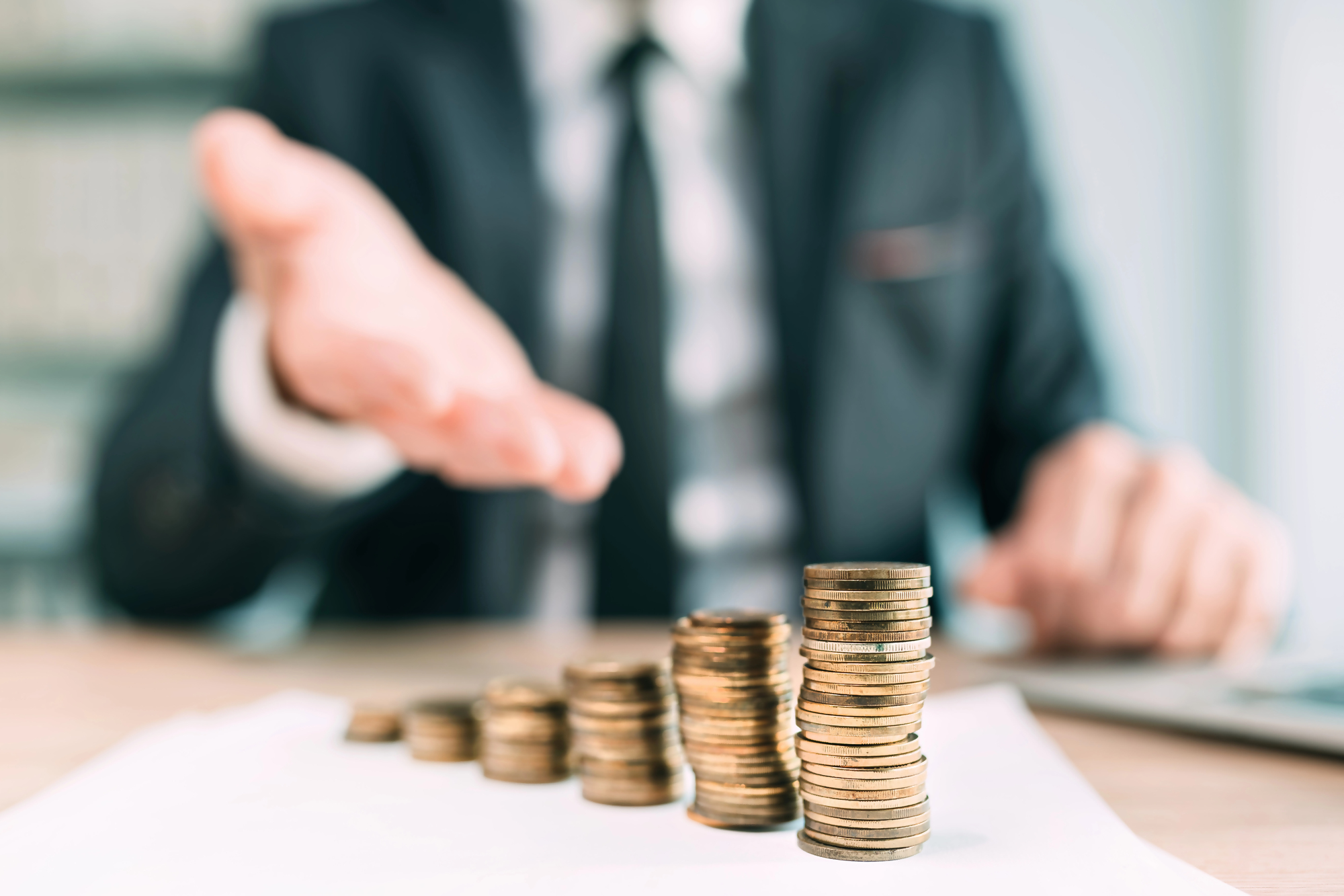 Pełnomocnictwo do konta bankowego – biznesmen w garniturze pokazuje na stosy monet ułożonych na biurku, symbolizując zarządzanie finansami i upoważnienie do dysponowania środkami na rachunku bankowym.