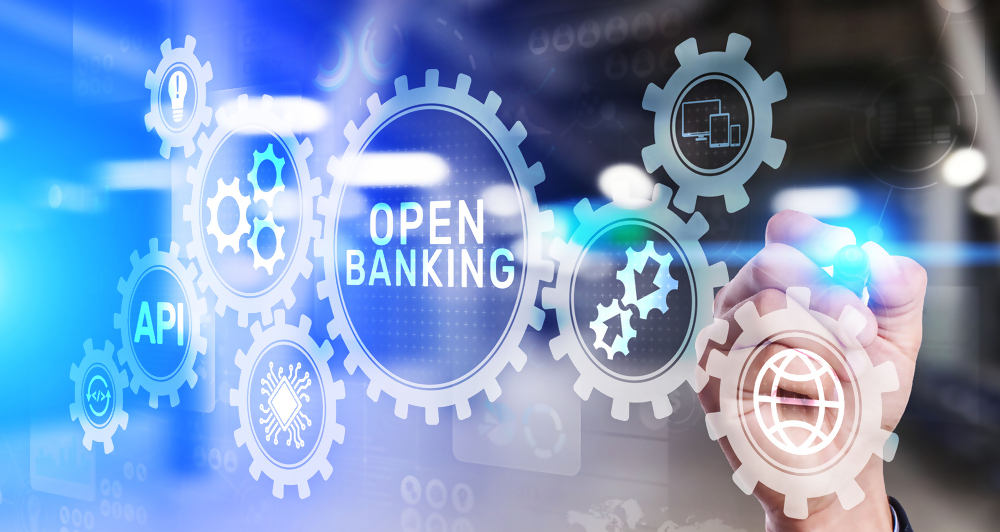 Grafika przedstawiająca koncepcję open banking, zębatki z napisami API i open banking, biznesowa dłoń rysująca schemat bankowości otwartej, motyw cyfrowy, nowoczesne technologie finansowe.