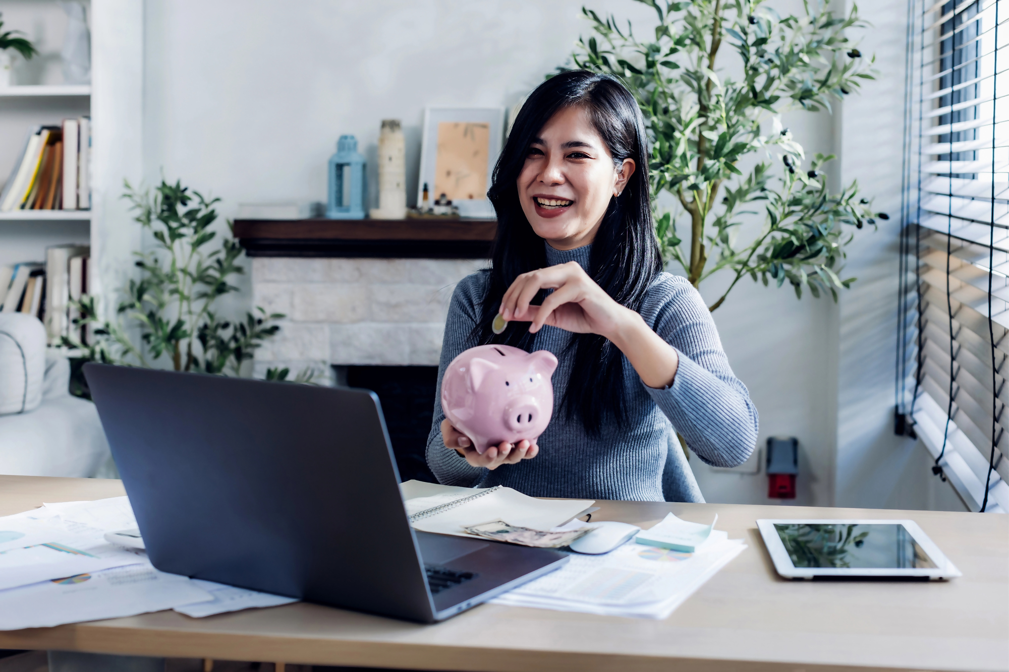 Kobieta trzymająca skarbonkę i odkładająca oszczędności przy komputerze, na biurku dokumenty finansowe związane z inwestowaniem w obligacje korporacyjne.