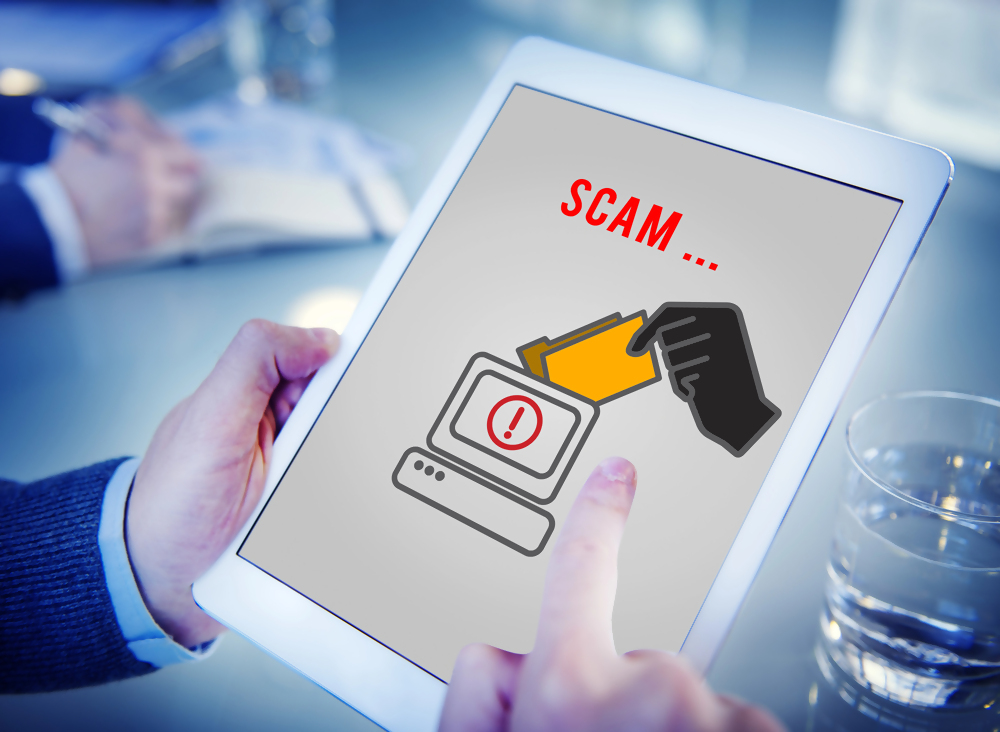 Osoba trzymająca tablet z wyświetlonym ostrzeżeniem o oszustwie internetowym, grafika z symbolem komputera, ręką sięgającą po dokument i napisem "scam", temat cyberbezpieczeństwa i zagrożeń online.